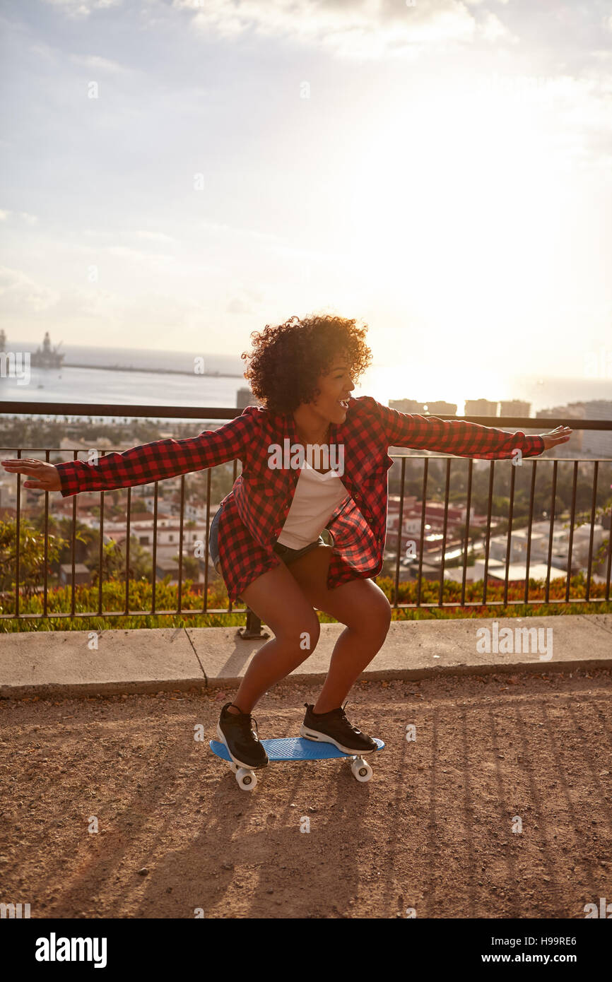 Skateboarding girl with her arms out and knees bent, on a bridge overlooking a city with a bay further behind, wearing casual clothing Stock Photo