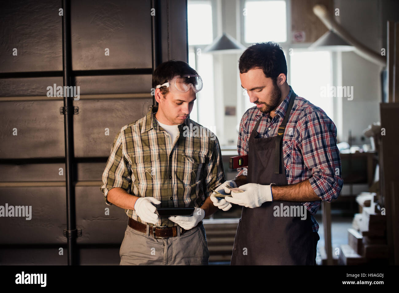Two carpenters are talking while holding papers and ruler. One of them is wearing safety mask. Carpentry shop background Stock Photo