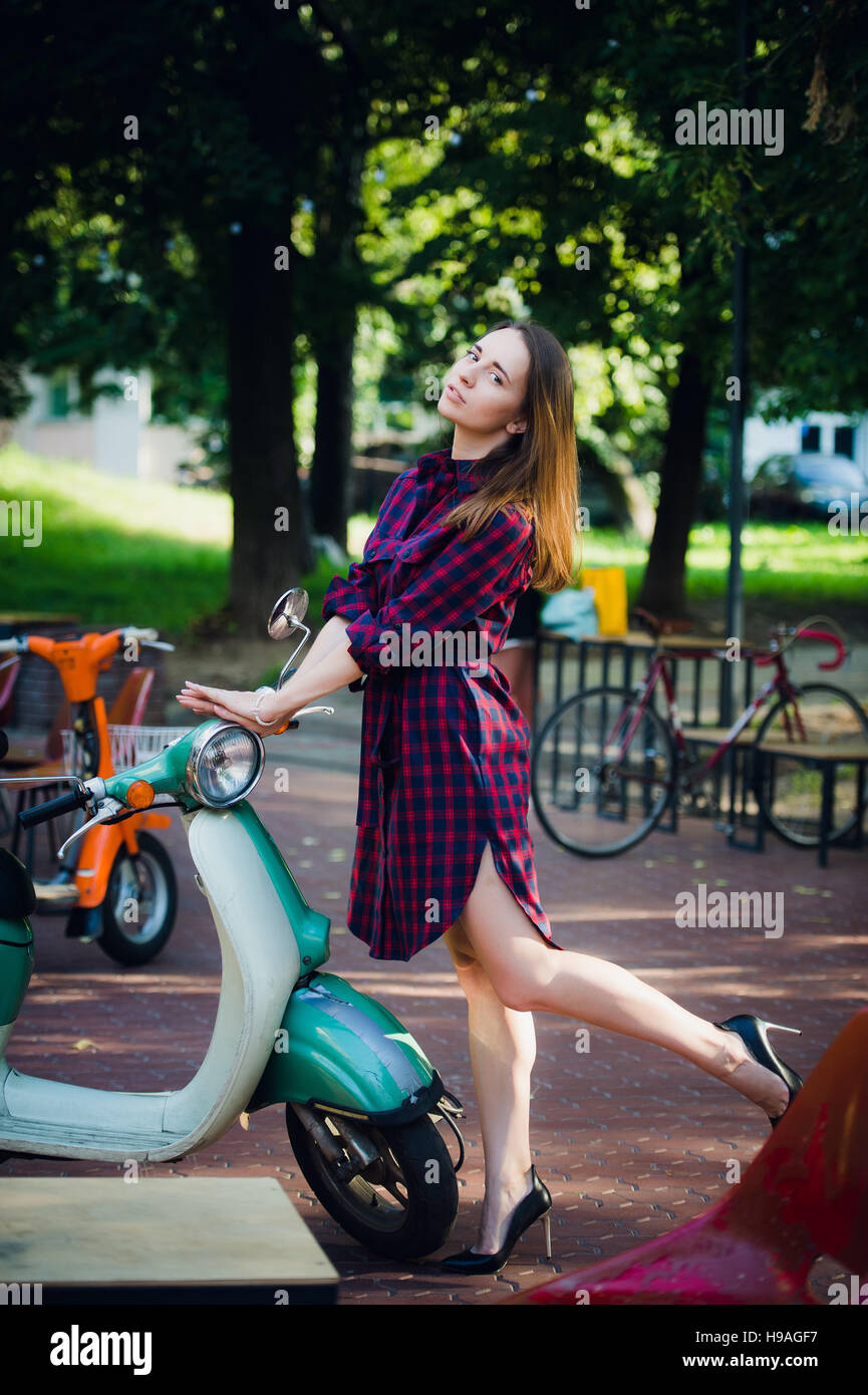 Urban fashion. Outdoor portrait of pretty young woman wearing checkered dress standing near scooter at park outdoors Stock Photo