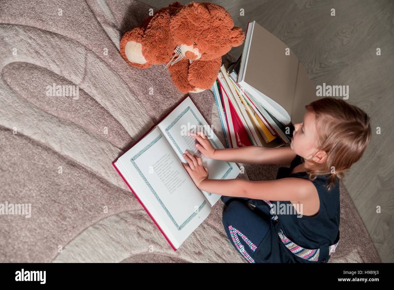 A little girl in a dark blue dress reading a book sitting on the floor ...