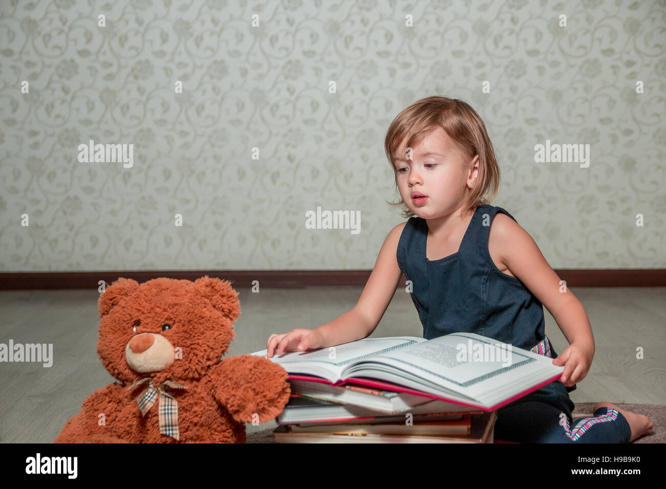 A little girl in a dark blue dress reading a book sitting on the floor ...
