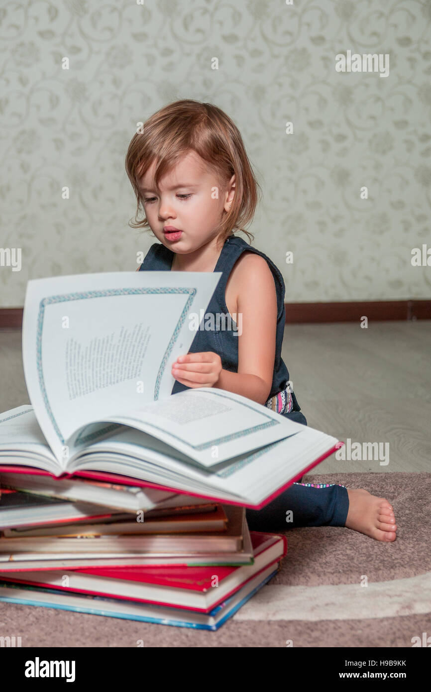 A little girl in a dark blue dress reading a book sitting on the floor ...