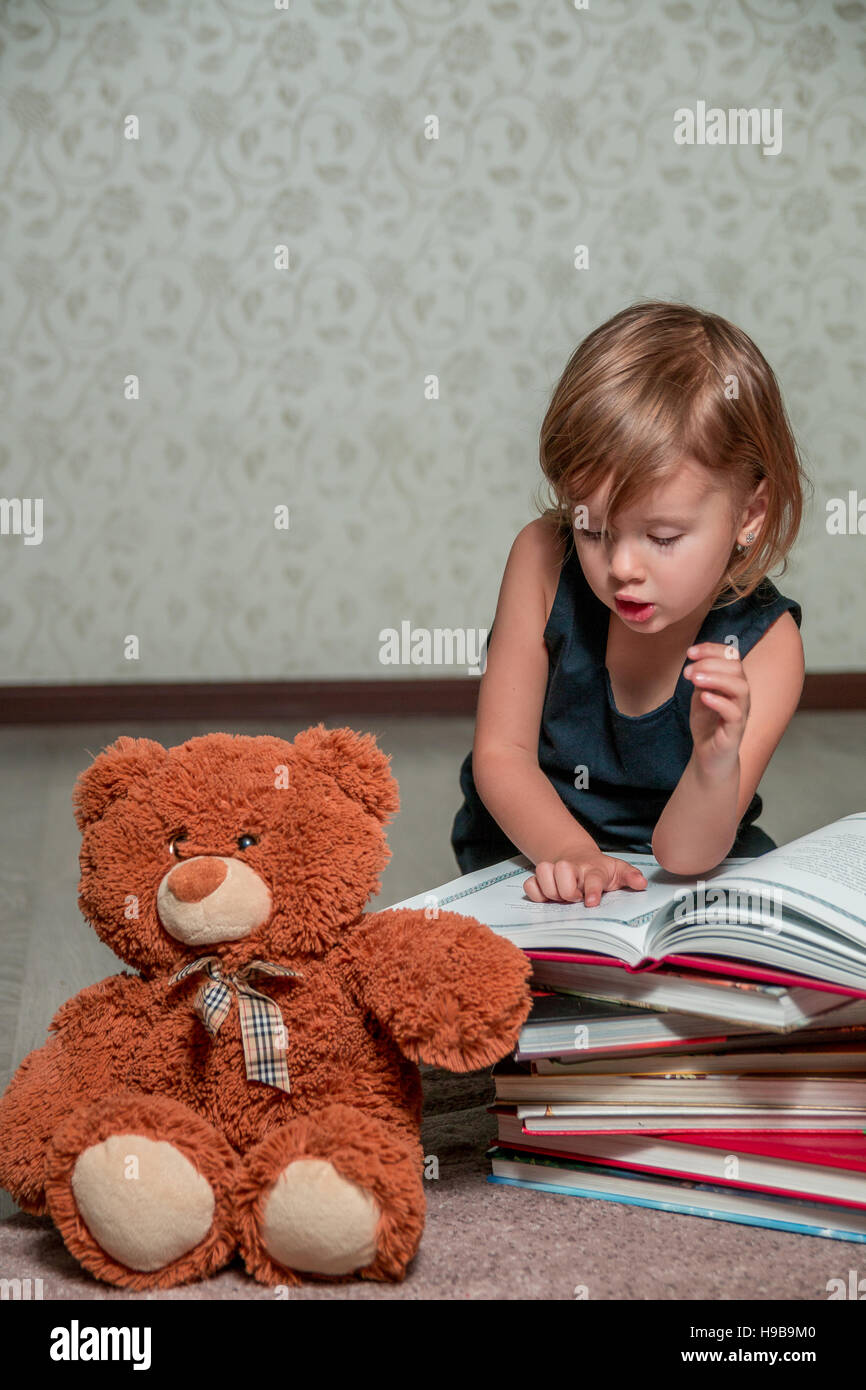 A little girl in a dark blue dress reading a book sitting on the floor ...