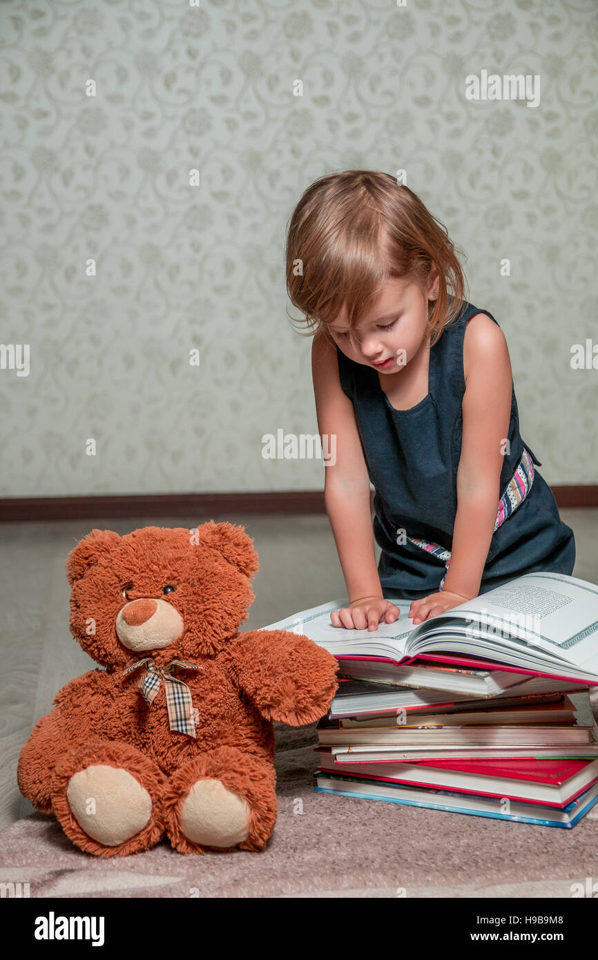 A little girl in a dark blue dress reading a book sitting on the floor ...