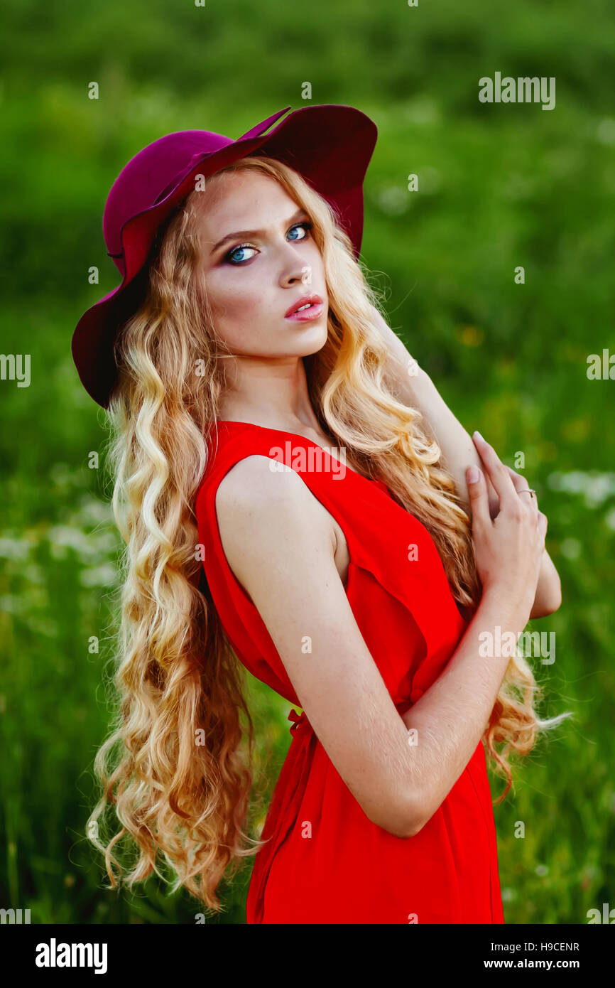 Blond girl with a long hair, in a red dress and red hat on a green background looking in camera Stock Photo
