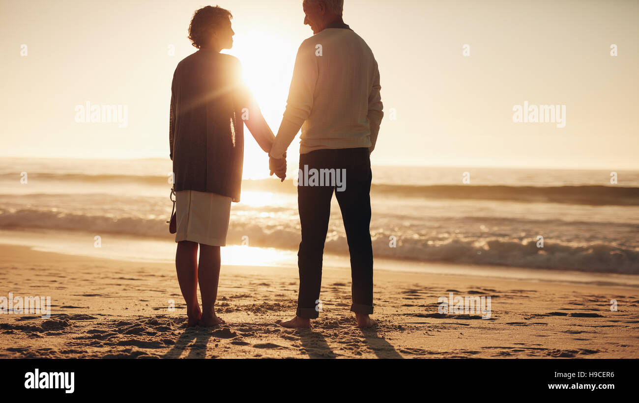 Rear view shot of a senior couple standing with holding hands and looking at each other while on the sea shore. Loving mature couple spending some tim Stock Photo