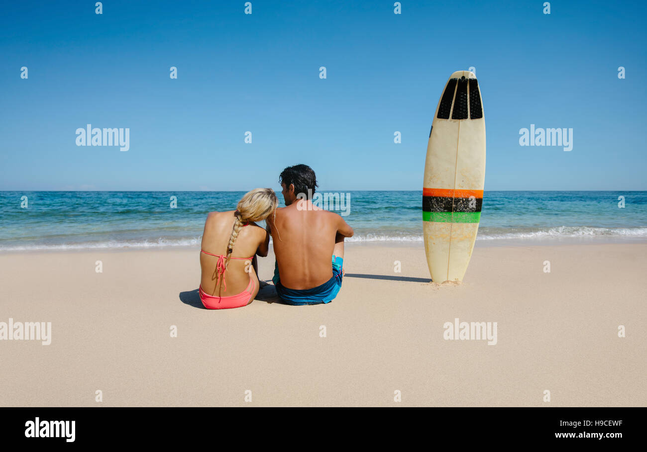 Rear view shot of young man and woman sitting on the beach with surf ...