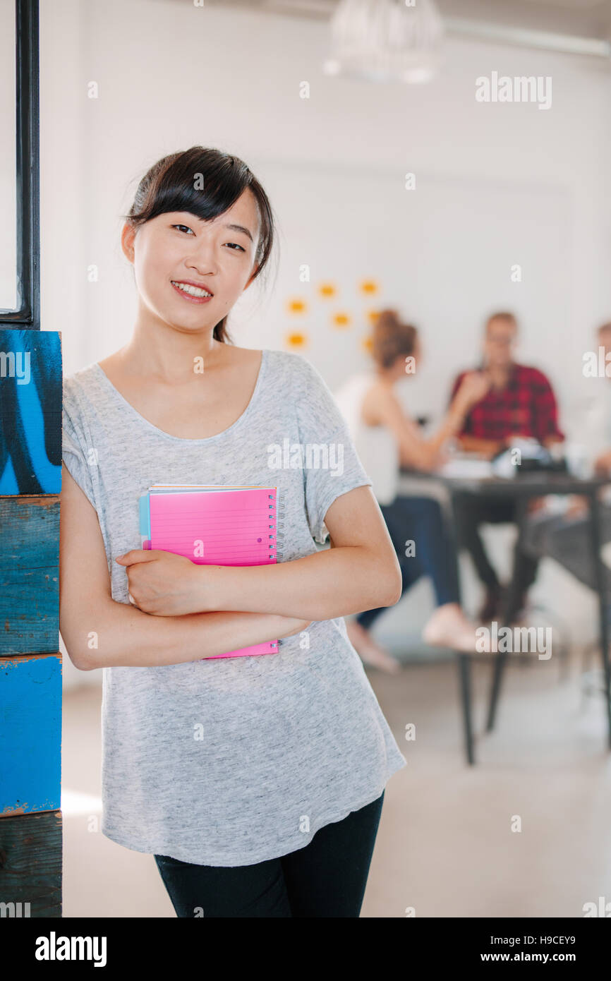 Portrait of a positive looking young business woman standing with her arms crossed with coworkers talking in the background. Stock Photo