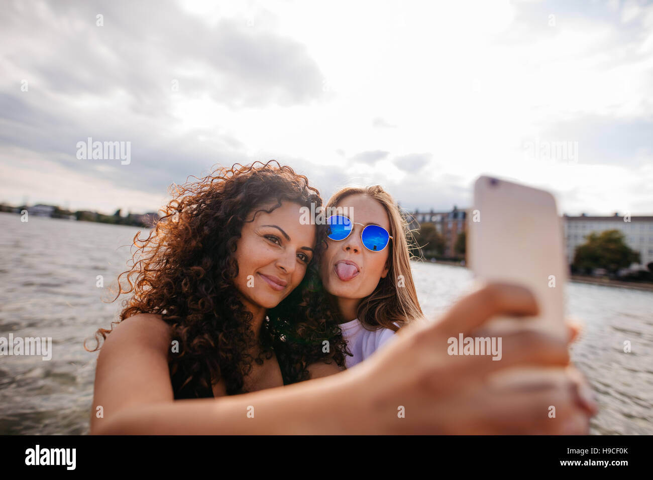 Shot of teenage girls taking selfie with smart phone by the lake. One sticking out tongue with other holding mobile phone. Stock Photo