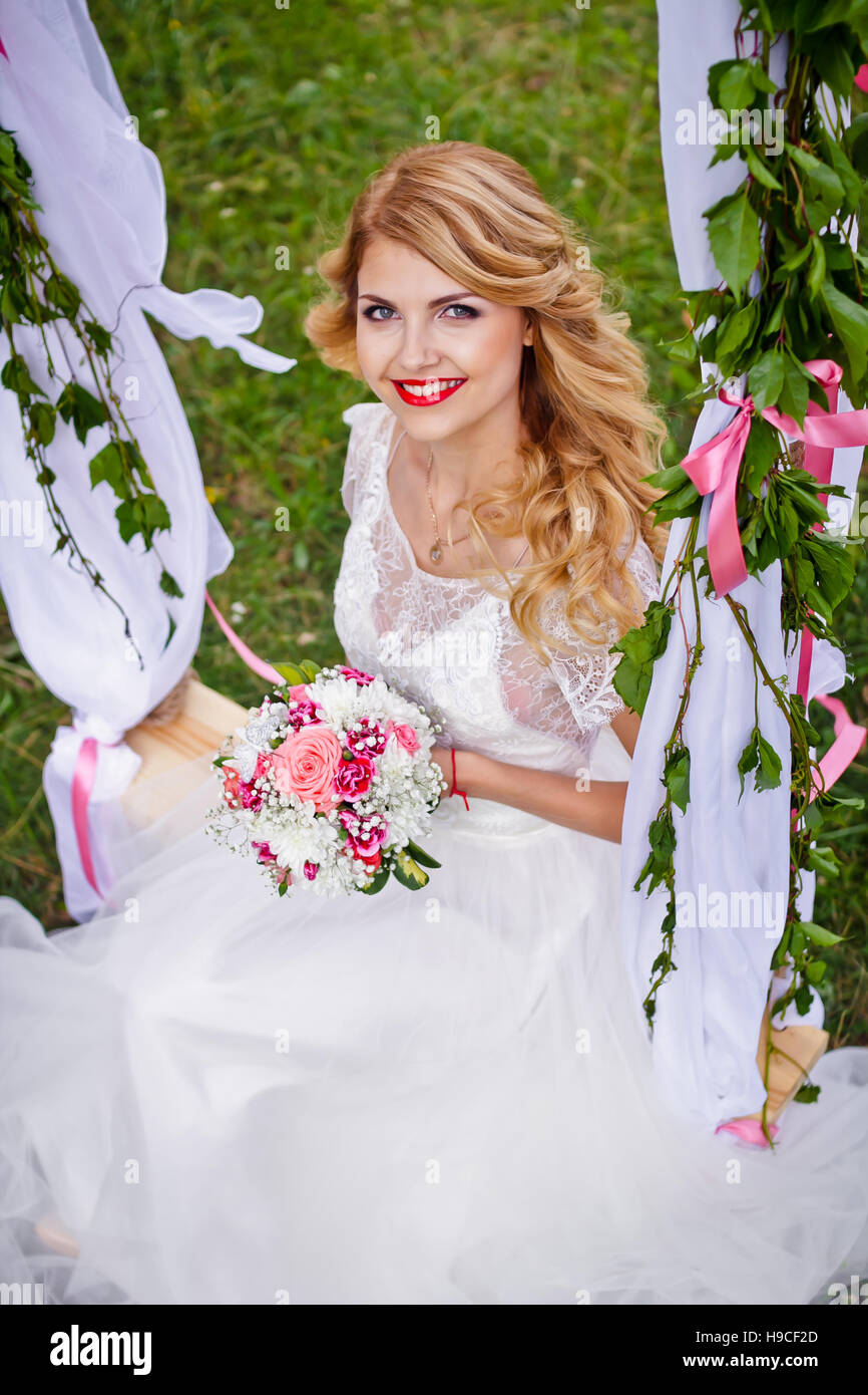 Beautiful blond bride in a white wedding dress swings on decorated swing and smiling to the camera Stock Photo