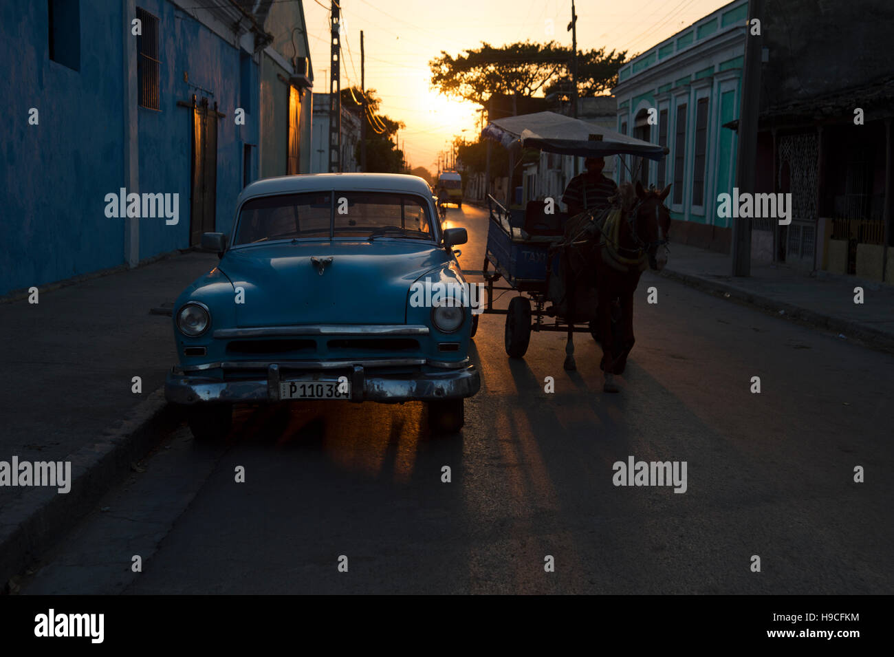 A horse and cart passes a classic US vintage car pared on a street in Cienfuegos Cuba with the sun setting behind Stock Photo