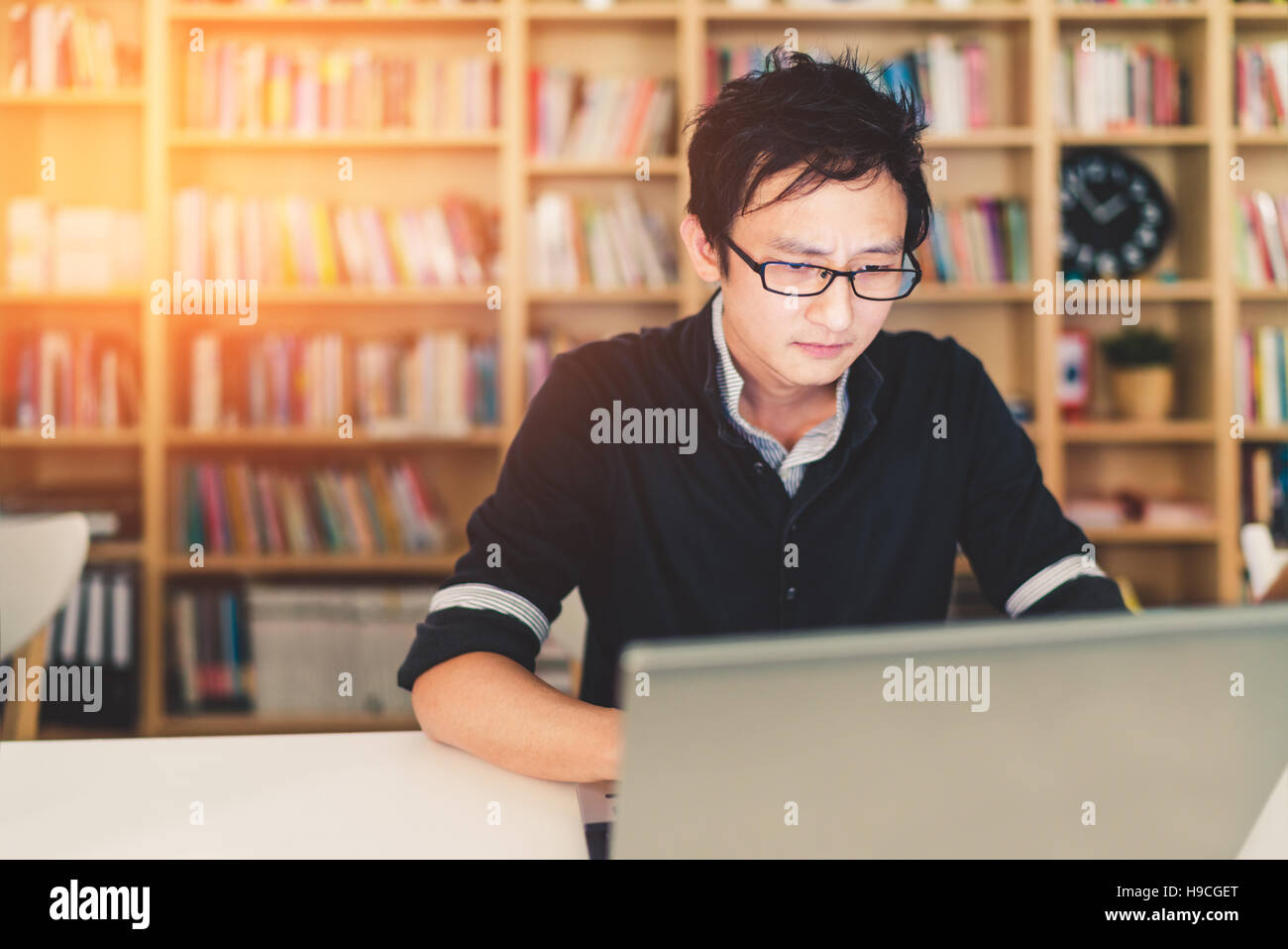 Young pensive Asian man working on laptop at home office or library with serious face, bookshelf blur background with copy space Stock Photo