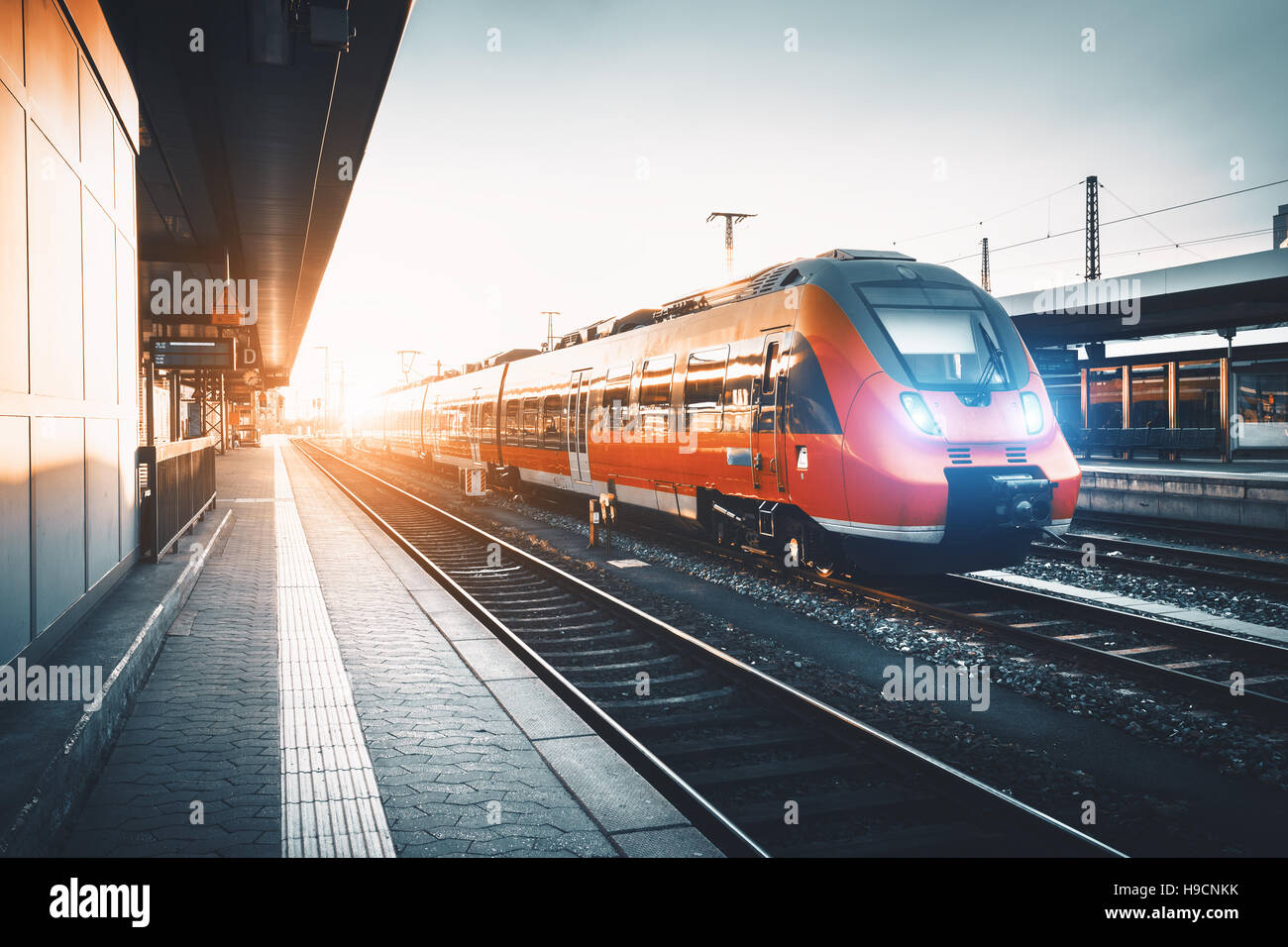 Modern high speed red commuter train at the railway station at sunset. Turning on train headlights. Railroad with vintage toning Stock Photo