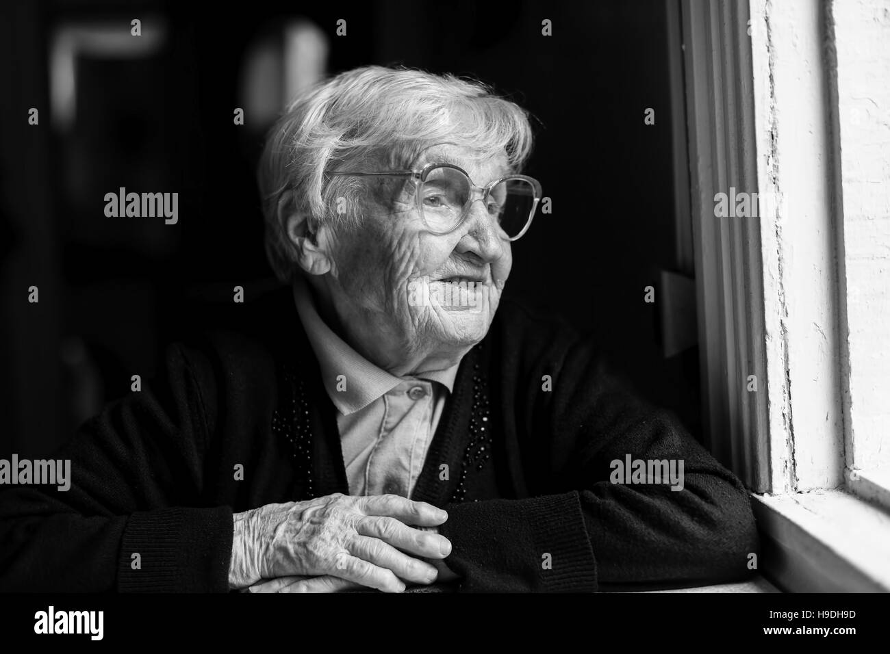 Old woman in glasses sitting by the window in the house. Stock Photo