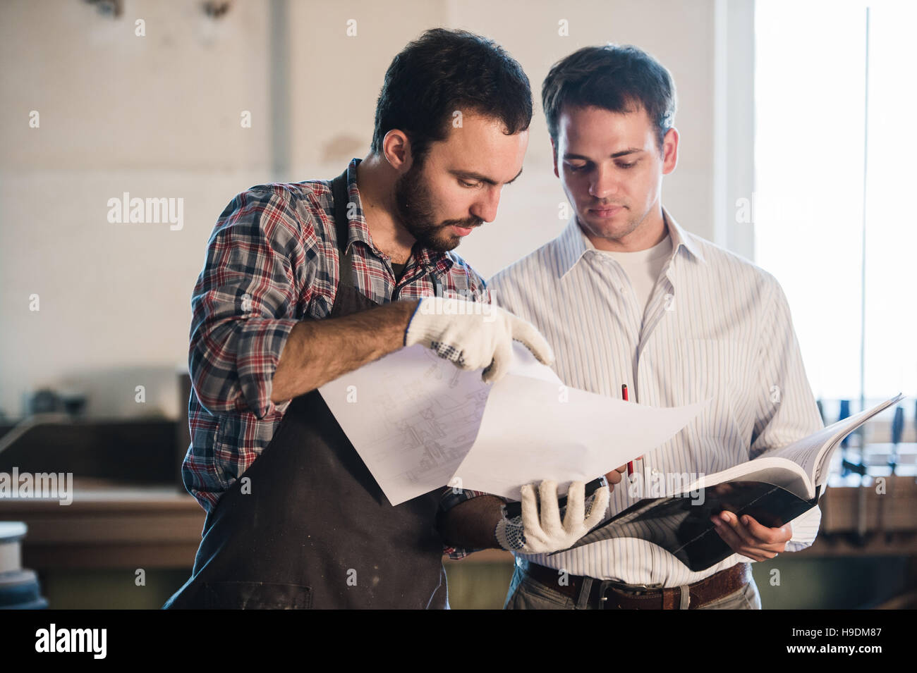 Happy male carpenter showing something to coworker at his notebook papers in workshop Stock Photo
