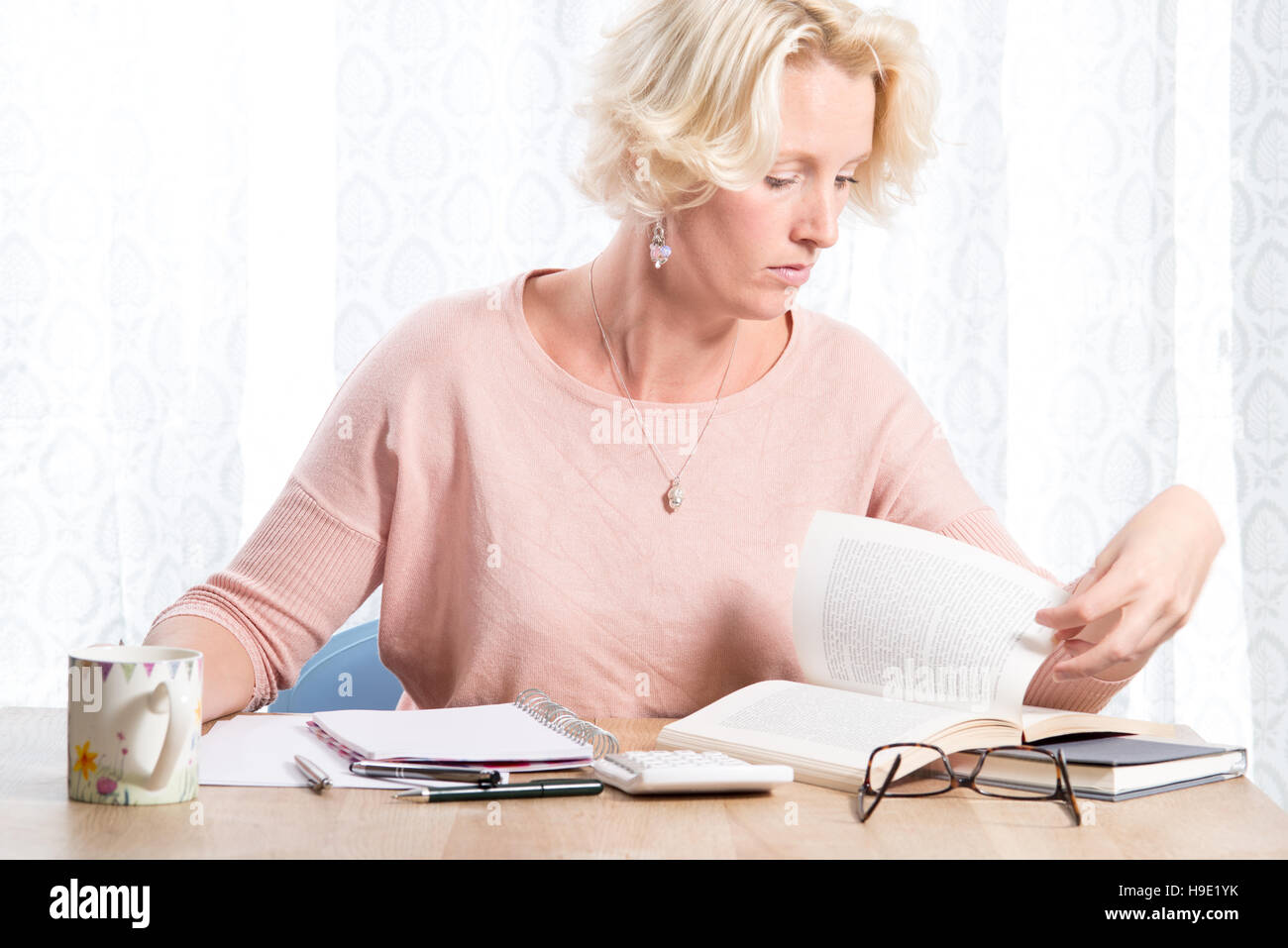 A blonde woman casually dressed in a pink jumper with necklace turns the page of a book while researching some work.  She sits at a wooden desk with a Stock Photo