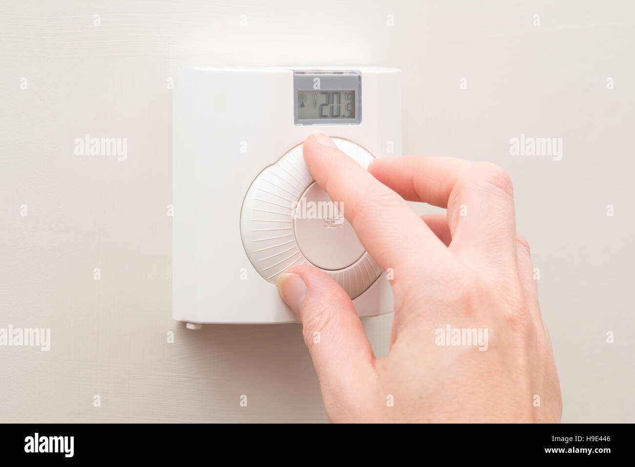 A hand turns the dial on a white wall mounted thermostat reading 20