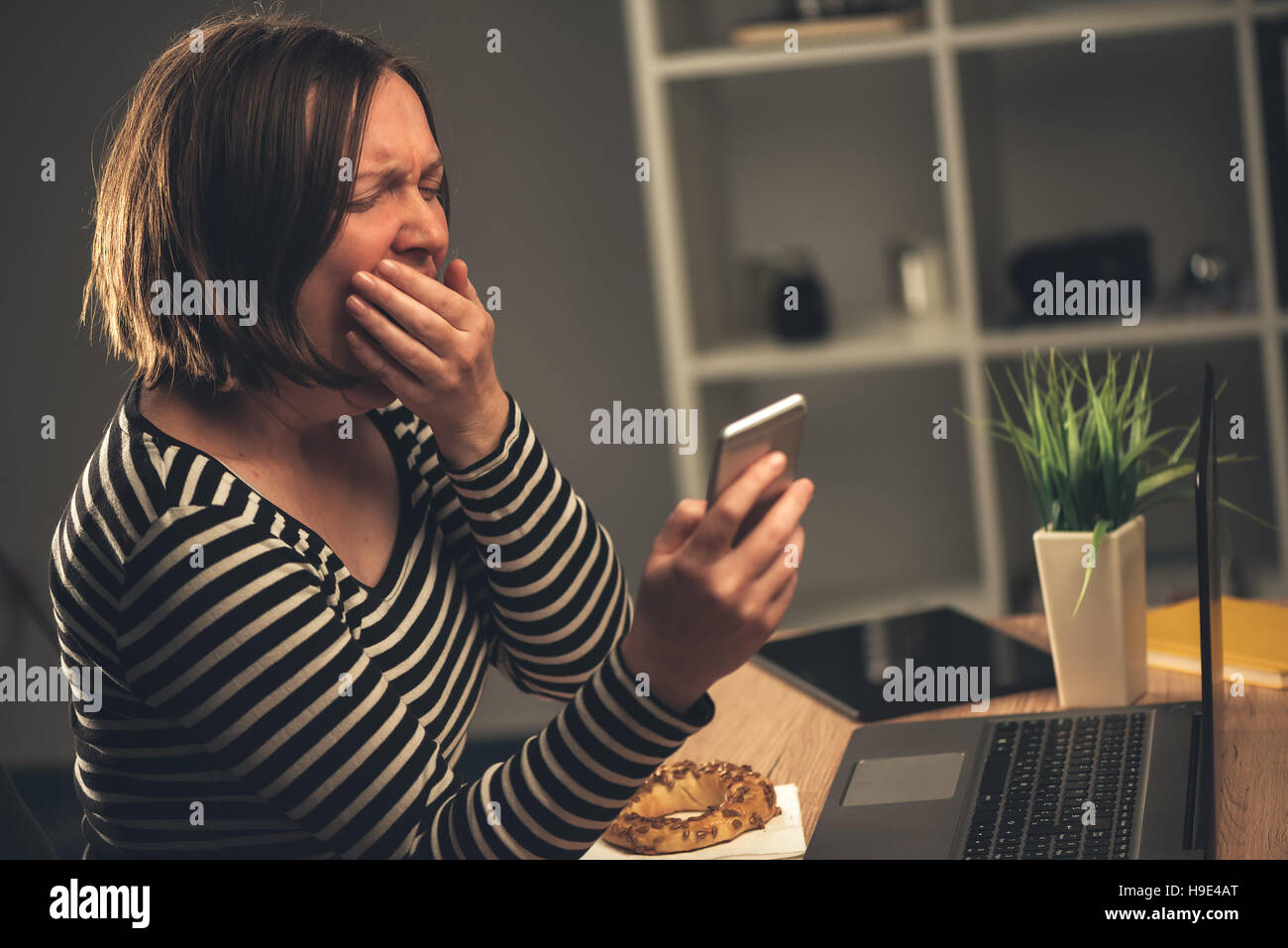 Tired businesswoman working overtime and yawning in office, female person answering to a text message on mobile phone Stock Photo