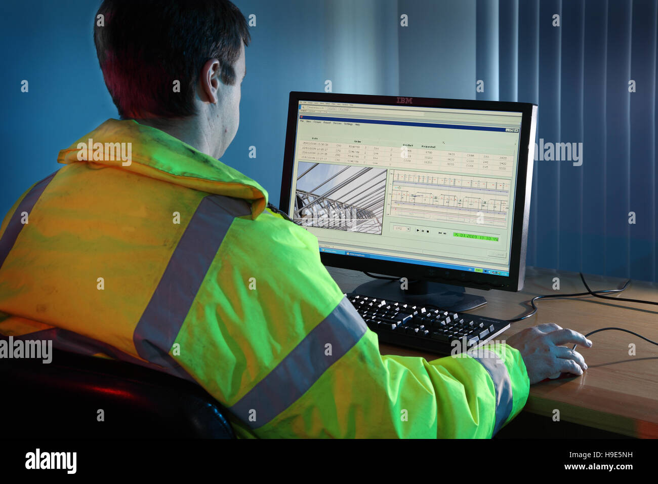 An engineer in a high-visibility jacket works at a computer in a construction site office. Screen shows structural analysis. Stock Photo