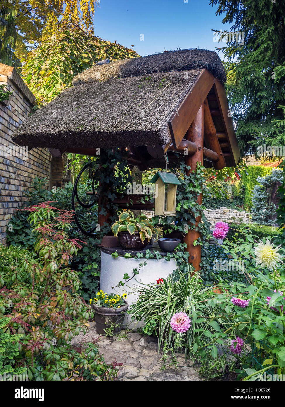 Old rustic thatched well in the garden surrounded by flowers and trees Stock Photo