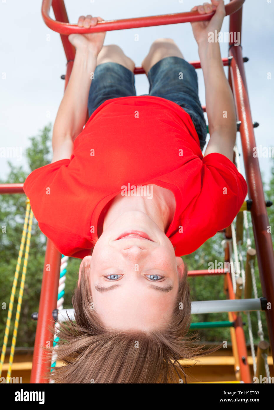 Low angle view of cute teen boy wearing red tshirt hanging upside down from a climbing frame in a playground looking at camera smiling Stock Photo