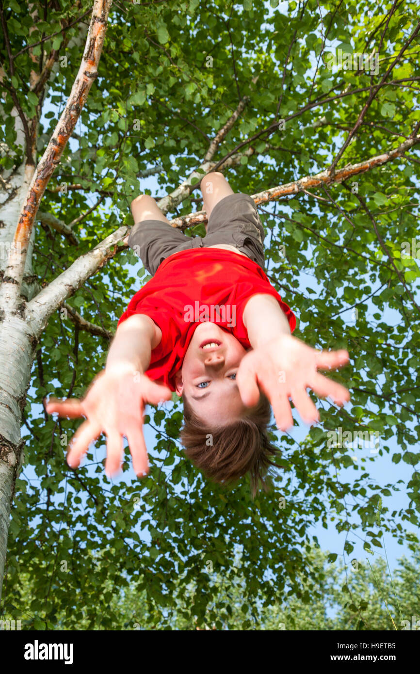 Low angle view of cute teen boy wearing red tshirt hanging upside down from a tree looking at camera with his hands and face blured in motion Stock Photo