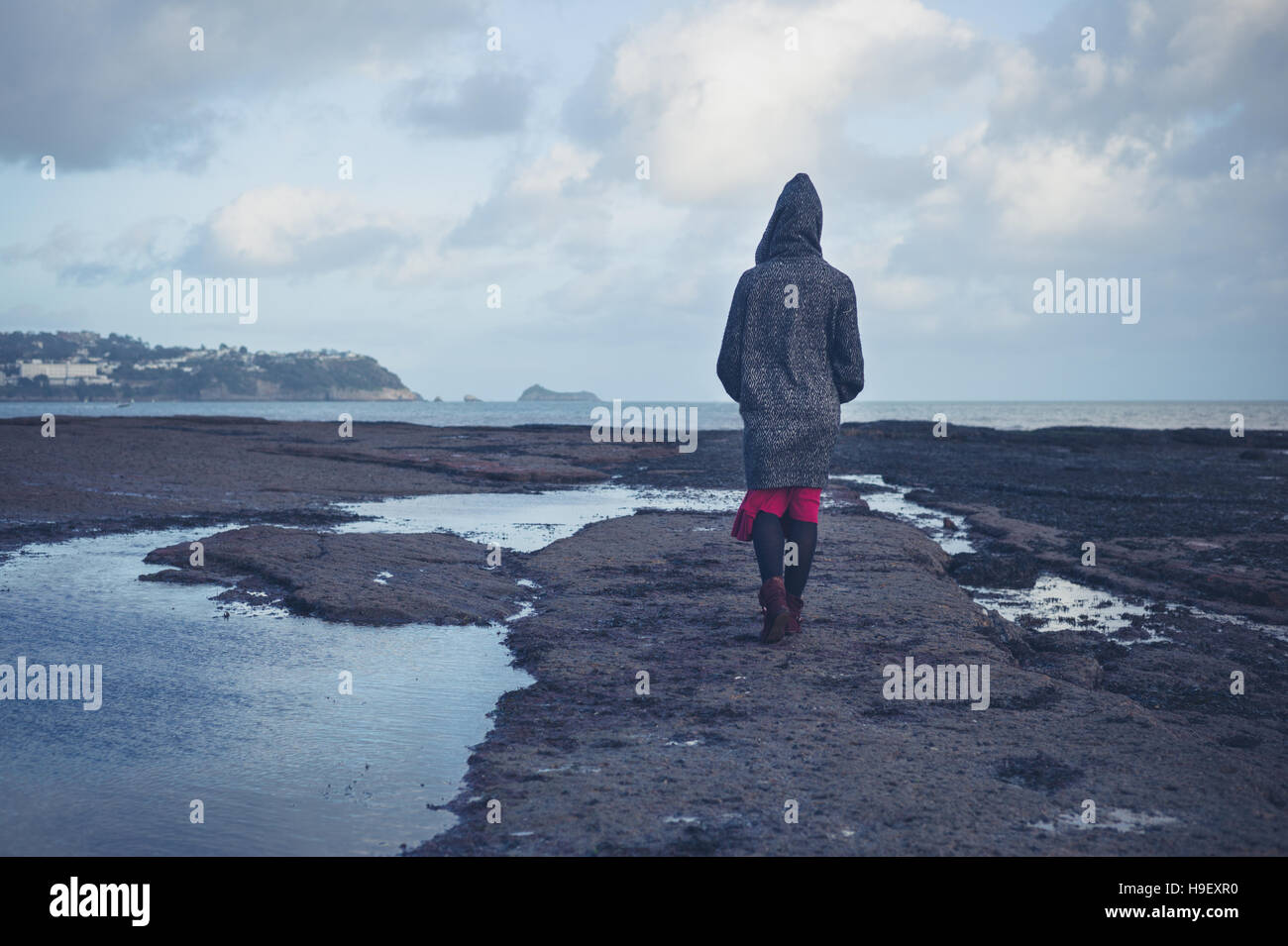 A young woman wearing a hooded coat is walking on a dramatic beach in ...