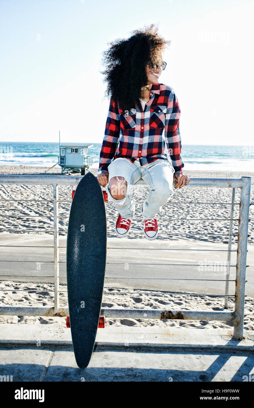 Hispanic woman sitting on fence at beach with skateboard Stock Photo