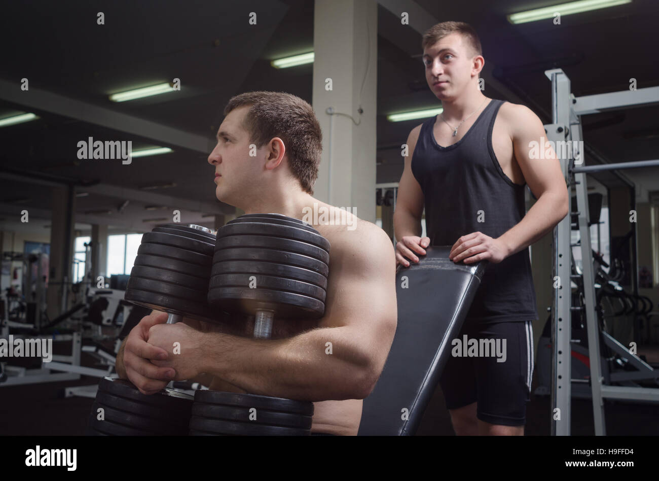 Two young muscular man preparing to bench press with dumbbells in the gym. A man with a dumbbell in the foreground, in the background a man for insura Stock Photo