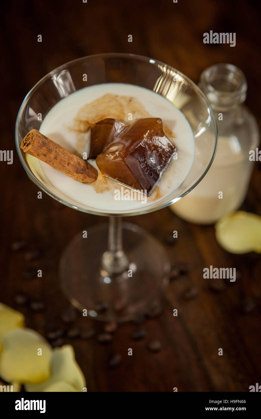 white cocktail poured over cinnamon stick and coffee ice cube in a martini glass on a wooden backdrop surrounded by rose petals Stock Photo
