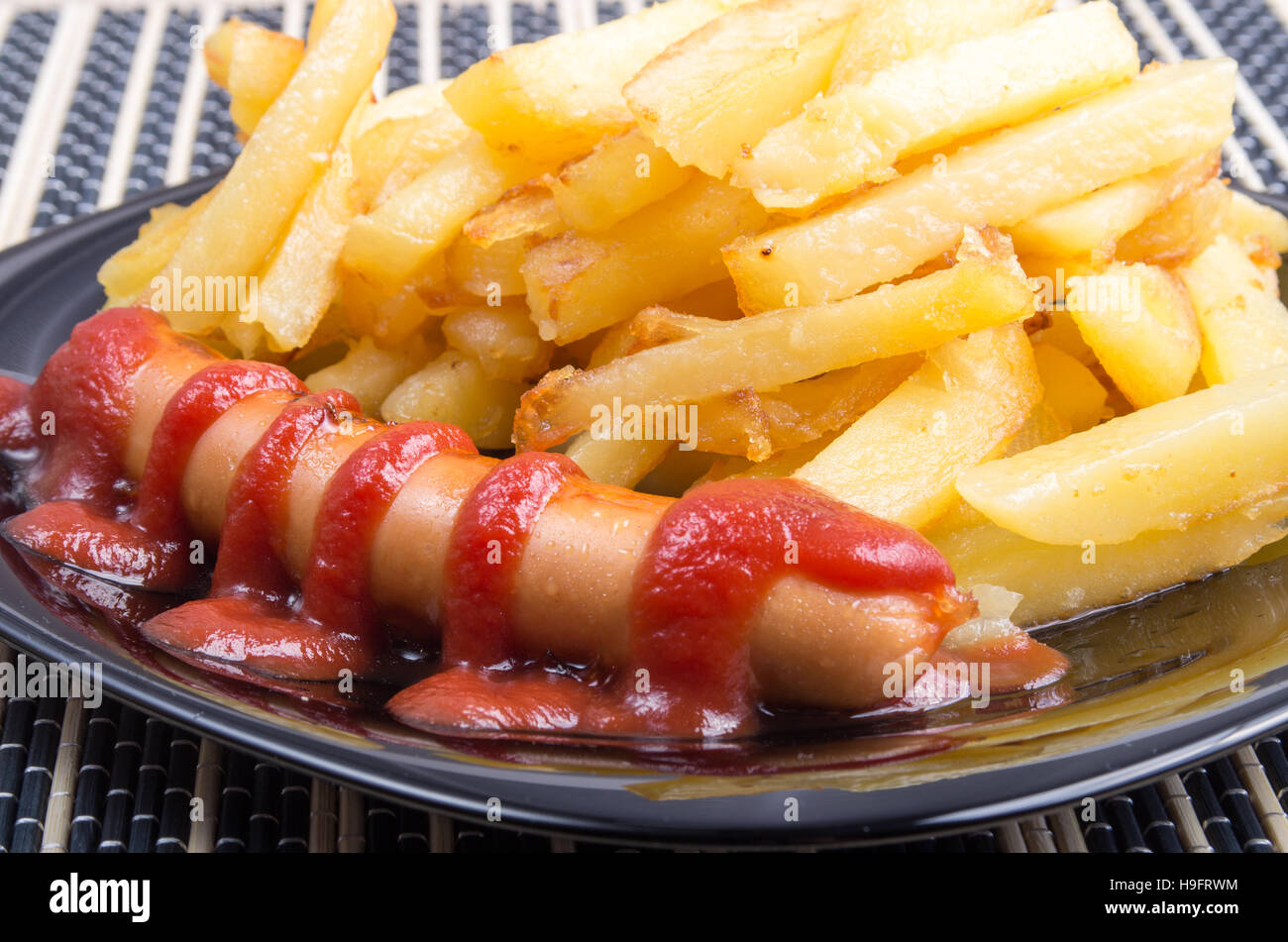 Fried sausage and french fries decorated with tomato sauce on a dark  plate close-up Stock Photo