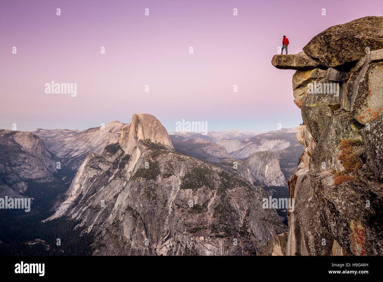 A fearless hiker is standing on overhanging rock at Glacier Point enjoying the view over Half Dome at sunset, Yosemite National Park, California, USA Stock Photo