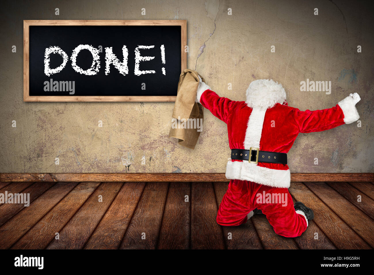 funny crazy hilarious red white santa claus celebration clench fist holding bag in the air job done blackboard on wooden floor interior room Stock Photo