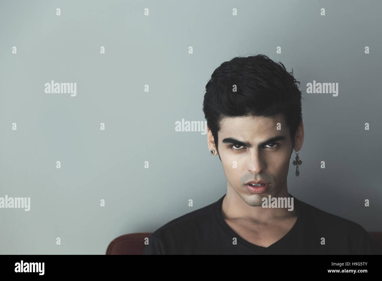 Portrait of attractive modern young white brazilian guy with black hair and cross-earring looking sullenly and saucily, gray wall behind Stock Photo