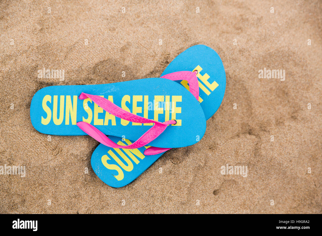 Pair of flip flops with 'Sun Sea Selfie' written on them on a sandy beach. Stock Photo