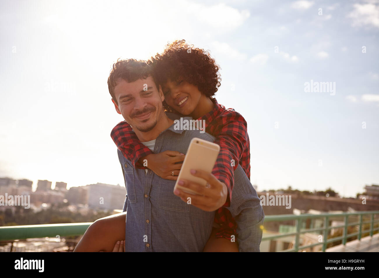 Laughing piggy back riding girl taking a selfie of them while sitting on her boyfriend back holding on with her right hand Stock Photo