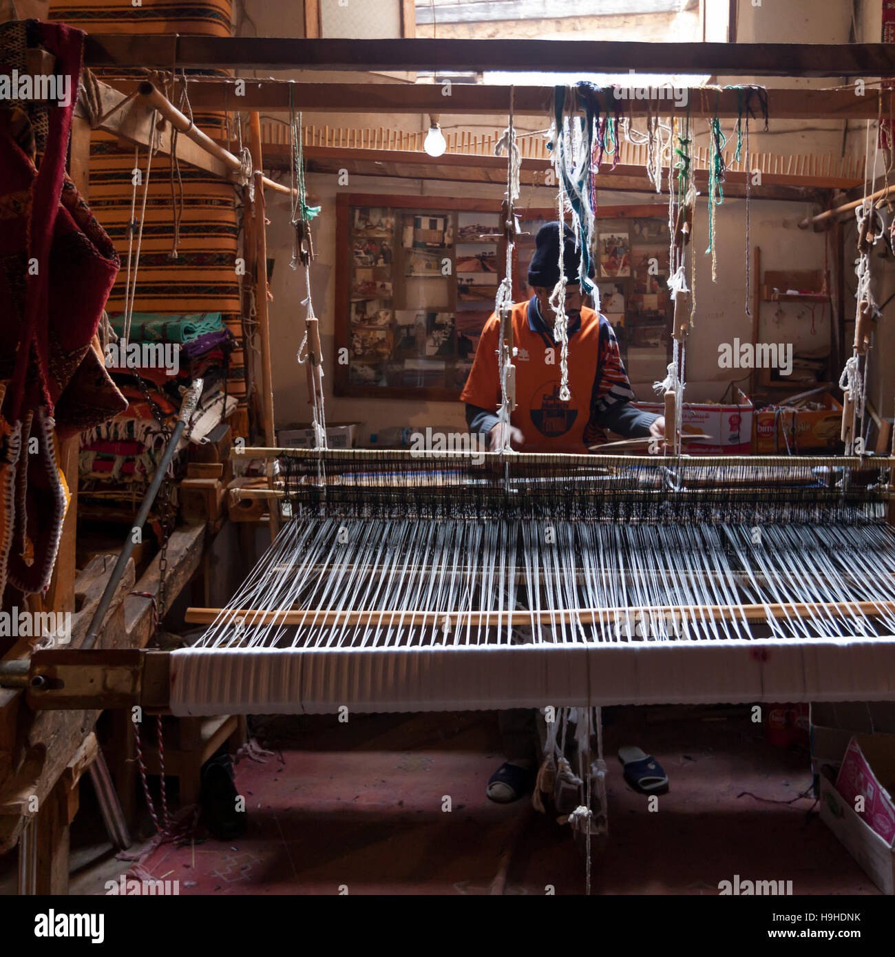 A weaver operates a wide loom in a workshop in Fès, Morocco, colourful fabric rolls and carpets hang on the wall behind him. Stock Photo