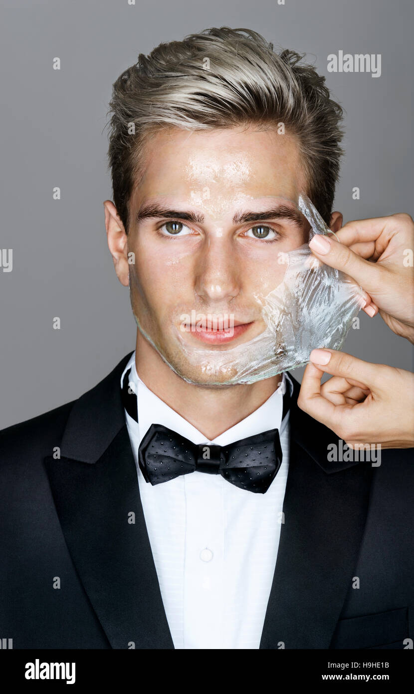 Portrait of well dressed elegant man in facial mask on gray background. Women's hands beautician removes peeling off a facial mask from the man's face Stock Photo