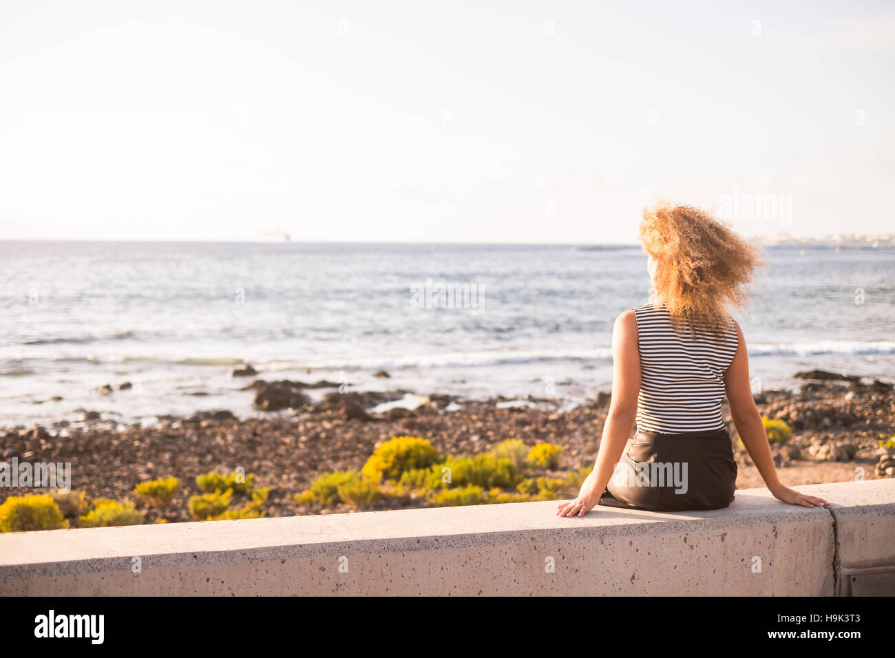 Back view of woman sitting on a wall looking at the sea Stock Photo - Alamy