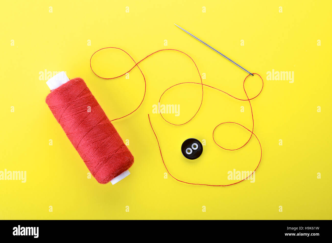 Spool of red thread, needle and clothing button on yellow background,  symbol of handmade and needlework. Stock Photo