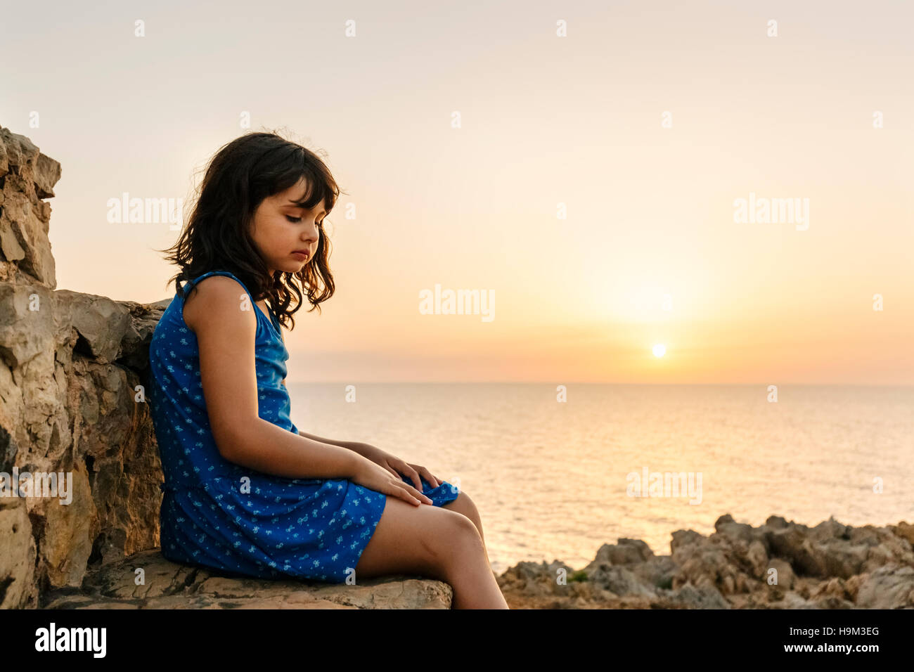 Sad little girl sitting in front of the sea at sunset Stock Photo - Alamy