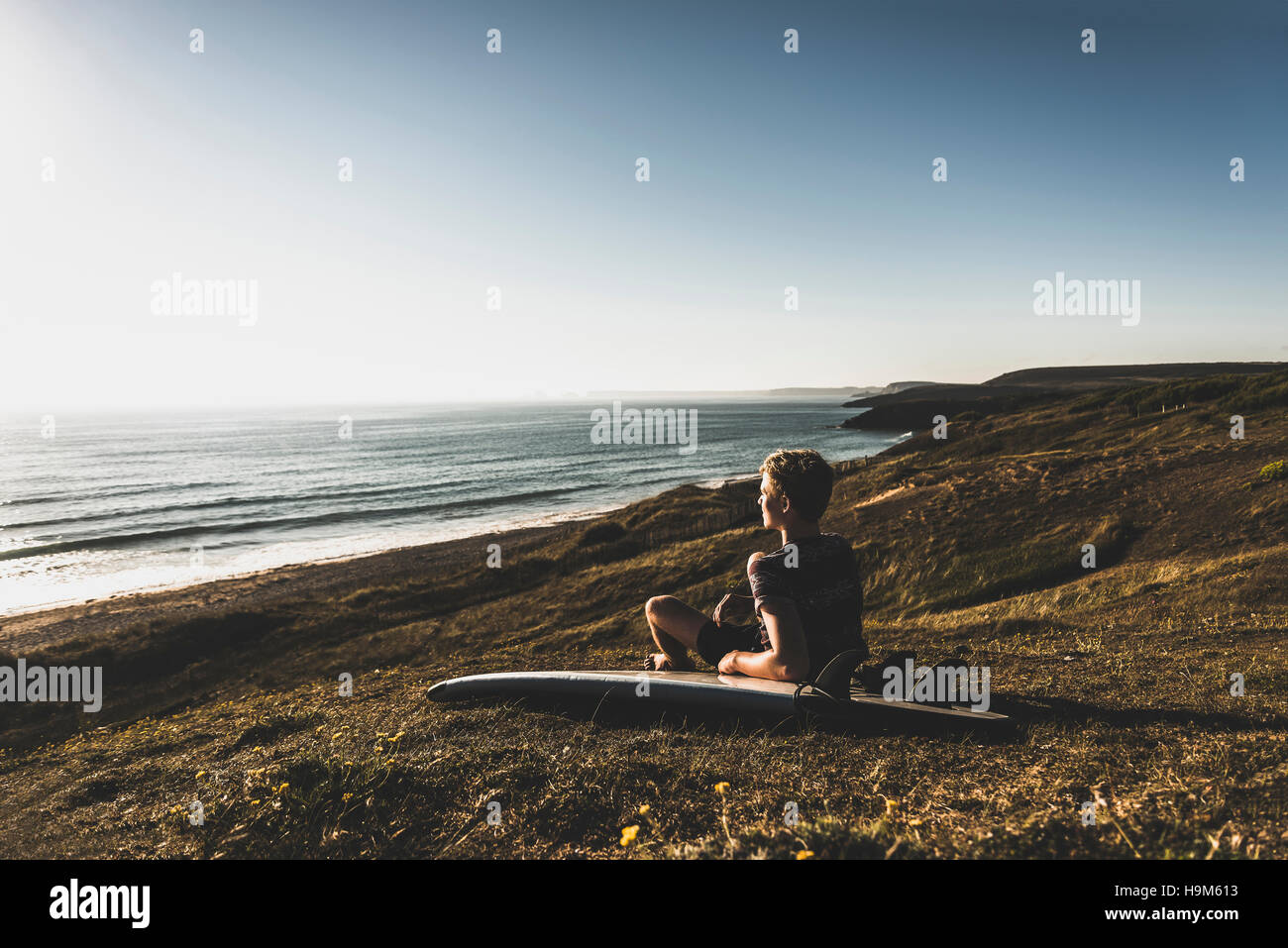 Teenage boy with surfboard sitting on the beach looking to the sea Stock Photo