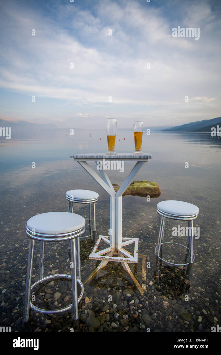 Color image of three chairs and a table with two beers, on the shores of a lake. Stock Photo