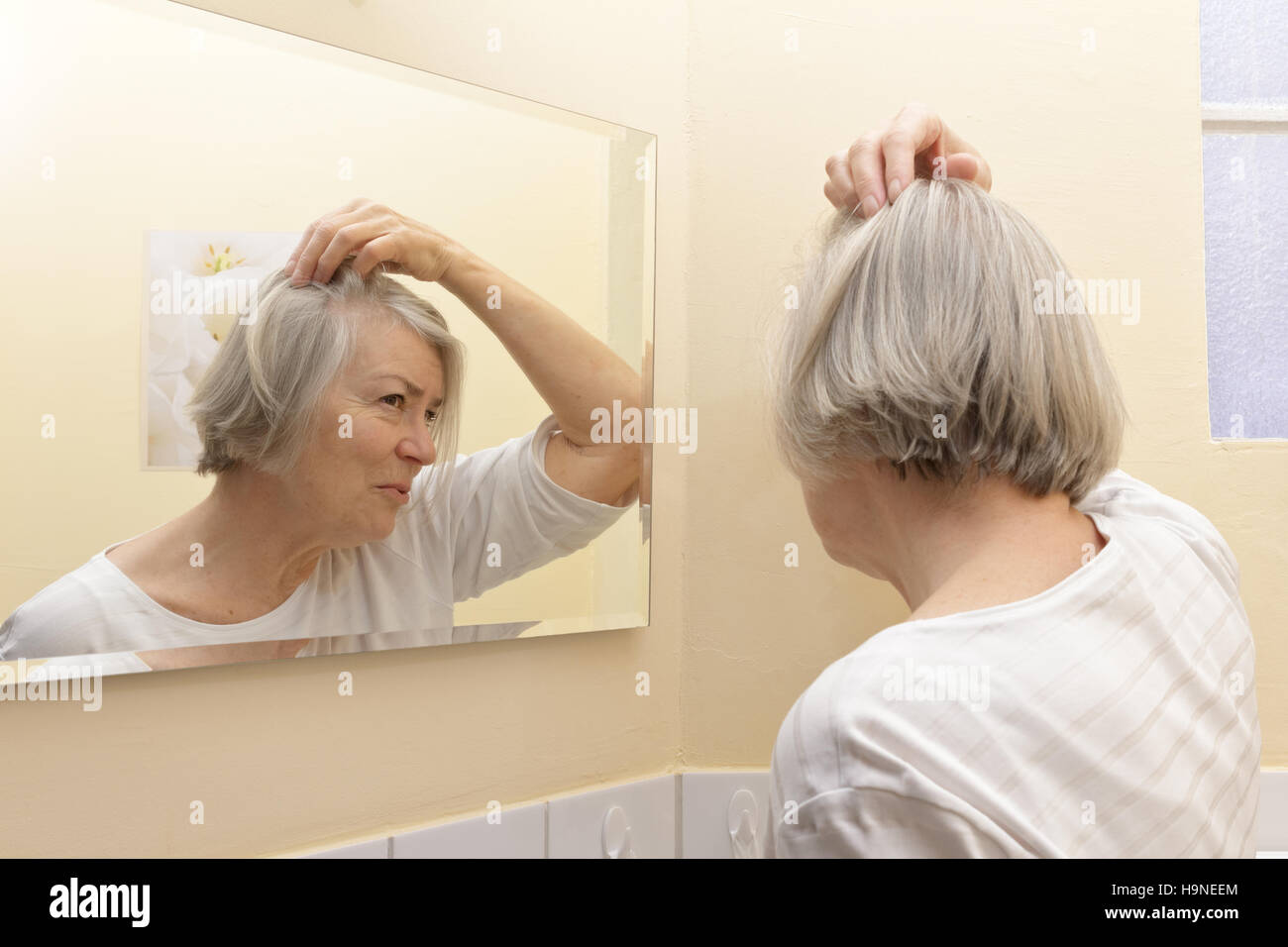Old woman with thin gray hair and a worried look on her face  examining her beginning baldness  in the mirror of her bathroom Stock Photo