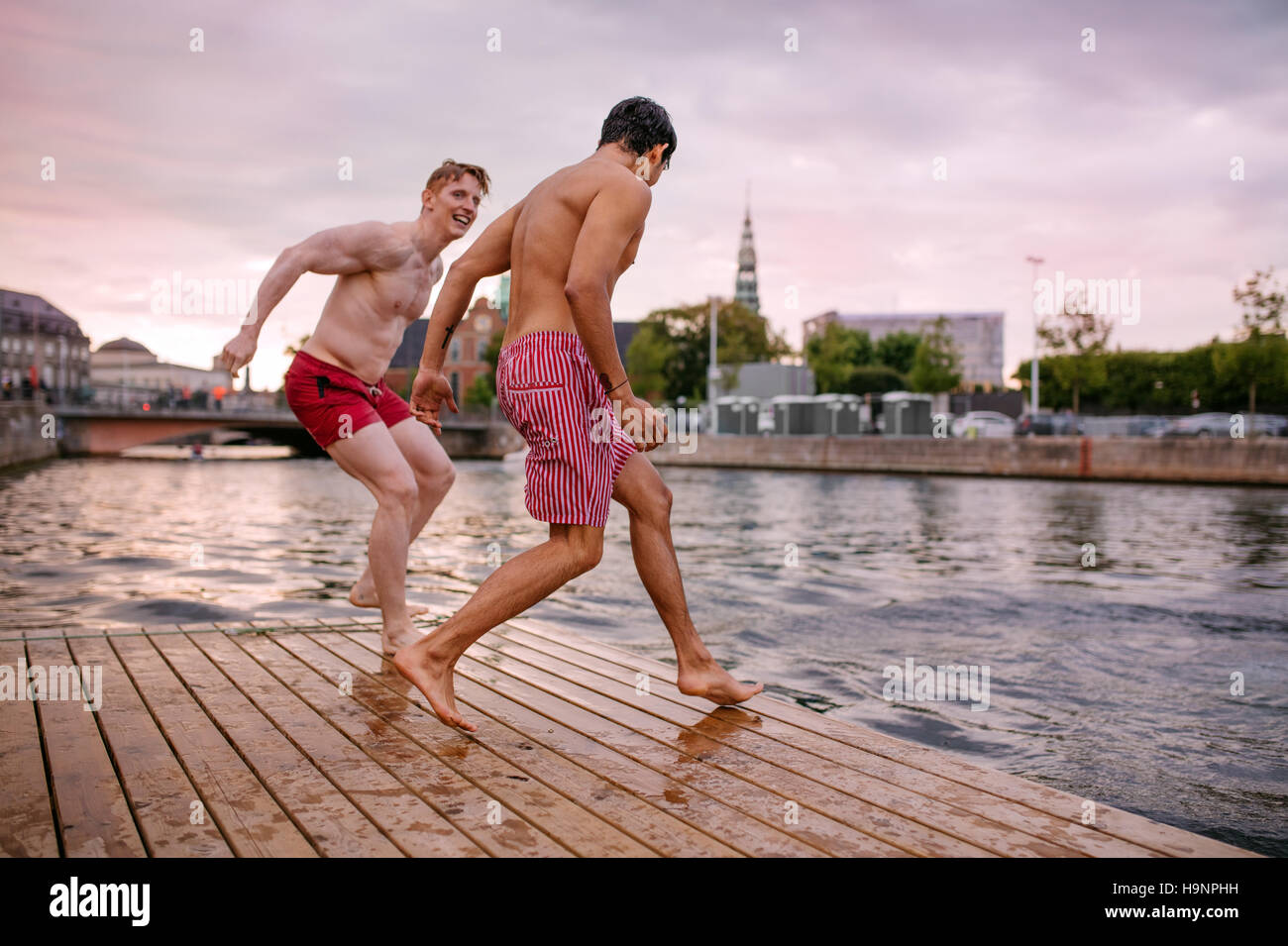Young friends about to jump into the lake. Young men running on a jetty. Stock Photo