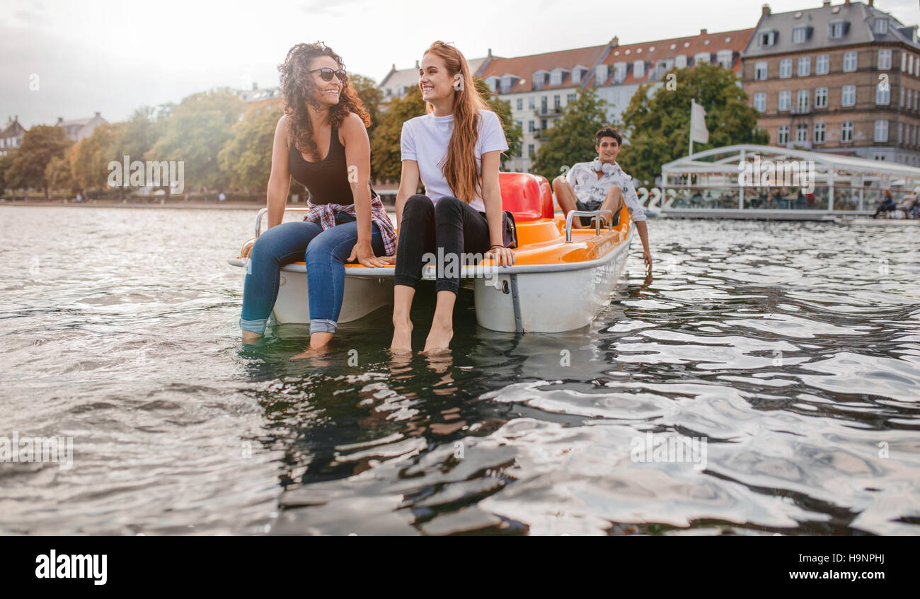 Outdoors shot of young female friends sitting in front pedal boat and putting their feet in water with man in background. Teenage friends enjoying boa Stock Photo