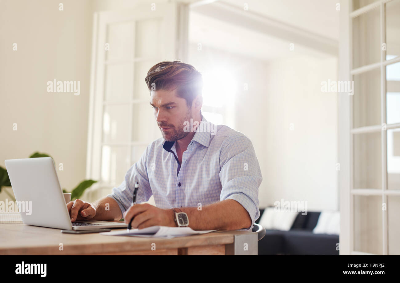 Indoor shot of young man working on laptop in the office and writing down notes. Business man working from home. Stock Photo