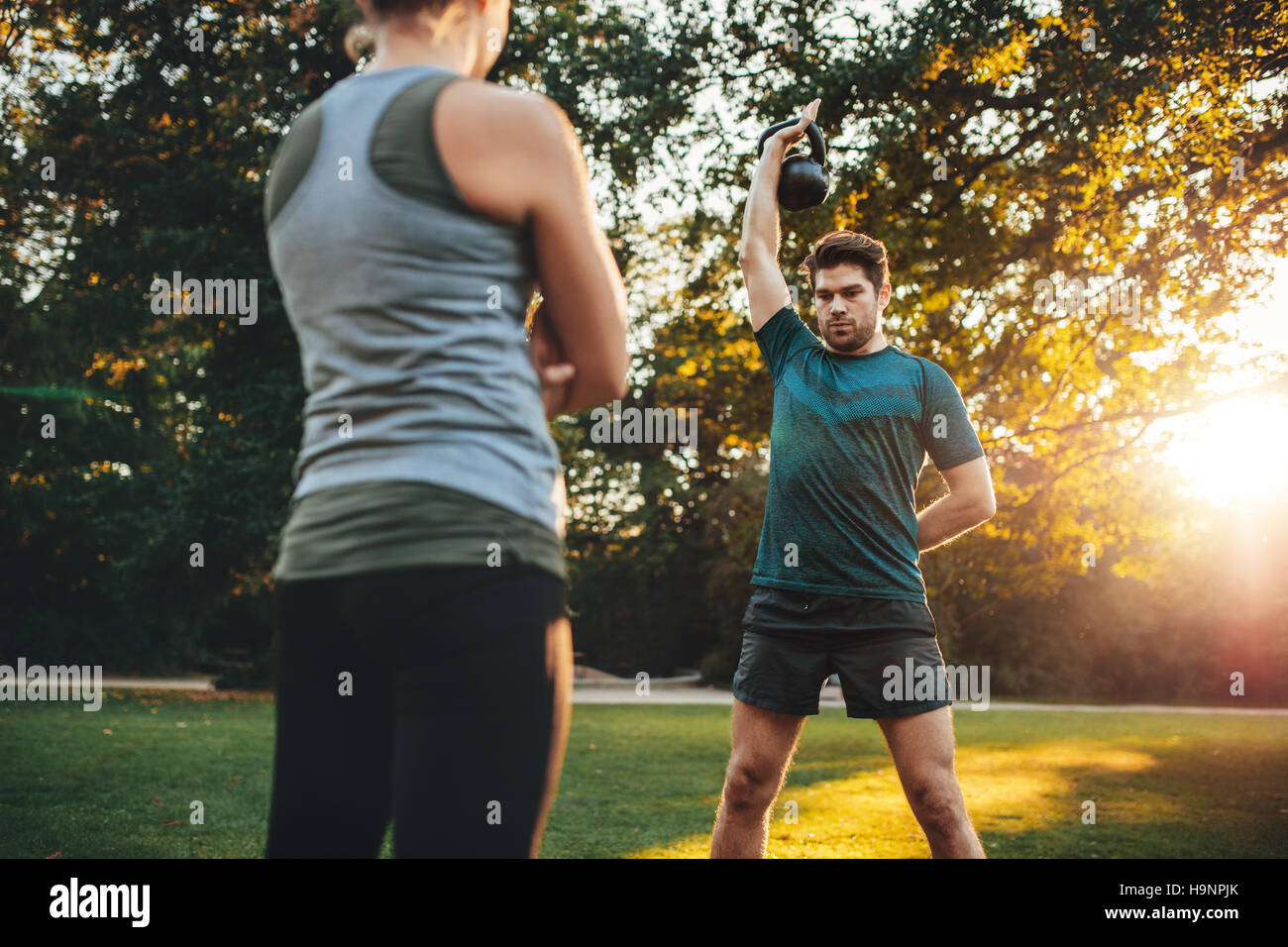 Young man doing weight training with personal female trainer in the park. Caucasian male exercising with kettlebell. Stock Photo