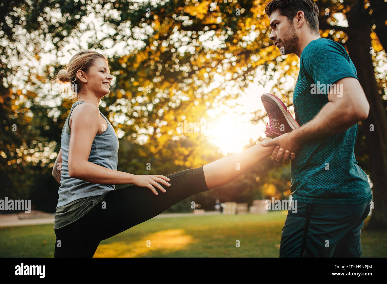 Personal trainer holding leg of woman stretching in park. Female exercising with support from her coach. Stock Photo