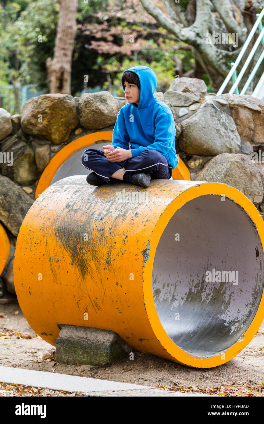 Japan, Akashi. Kazki, early teenage caucasian boy in blue hoodie sitting cross-legged on top of yellow tunnel section in play-park. Alone. Thoughtful. Stock Photo